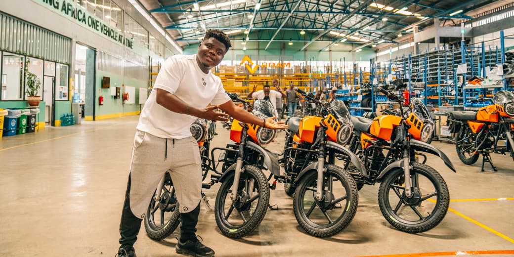 A man presenting a row of orange ROAM electric motorbikes inside the ROAM assembly facility. Credit: ROAM.